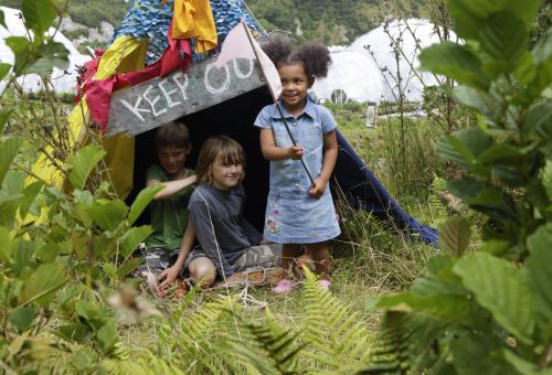 Three children sitting/standing by a handmade den onsite at the Eden Project
